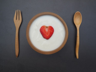 Red strawberry ripe piece slice of shape heart in yogert cream white color in a wood bowl with spoon and fork on the black cement background using for design banner healthy or diet food, See top view.