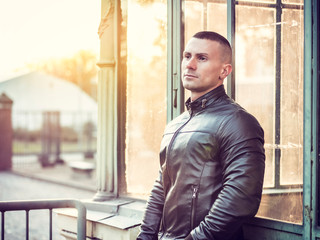 One handsome young man in urban setting in European city, standing, wearing black leather jacket and jeans at sunset