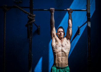 Blond, attractive young shirtless muscular man hanging from gym equipment, looking at camera