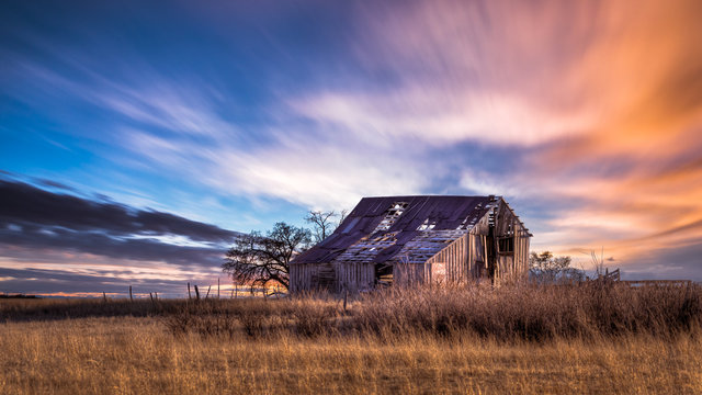 Old rural farmhouse in the countryside during a very colorful sunset. There is a grass meadow and a few trees.