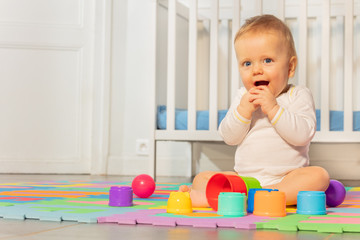 Beautiful baby boy at toddler age, hands near face sitting in front of crib play with toys on the carpet