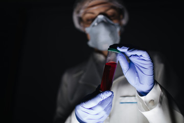 Medical specialist in protective uniform holding test tube with blood sample over dark studio background. Female expert holding glass container for testing in laboratory