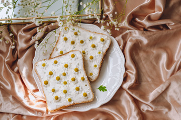 Toast bread sandwich with chamomile flowers in a white plate, on a golden satin fabric with gypsophila flowers. Spring morning