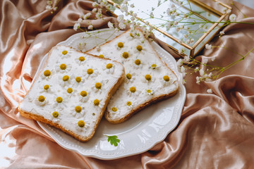 Toast bread sandwich with chamomile flowers in a white plate, on a golden satin fabric with gypsophila flowers. Spring morning