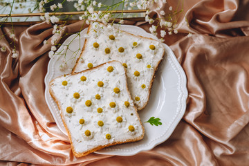 Toast bread sandwich with chamomile flowers in a white plate, on a golden satin fabric with gypsophila flowers. Spring morning