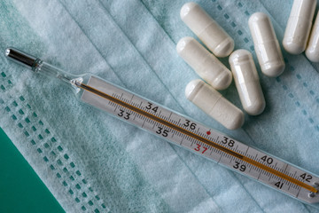 A mercury thermometer with a body temperature of 39 degrees on the scale, tablets and a disposable medical mask. Selective focus.
