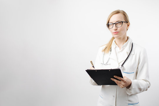 Thoughtful Mature Woman In Medical Clothing With Stethoscope On Neck Writing On Clipboard And Looking Aside. Female Doctor With Blond Hair Posing In Studio With White Background.