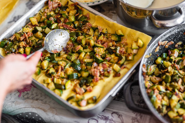Homemade preparation of lasagna with zucchini and bacon. Close up of female hand putting and spreading bechamel and courgettes cut into small pieces on lasagna. Selective focus.Pots, plates and trays