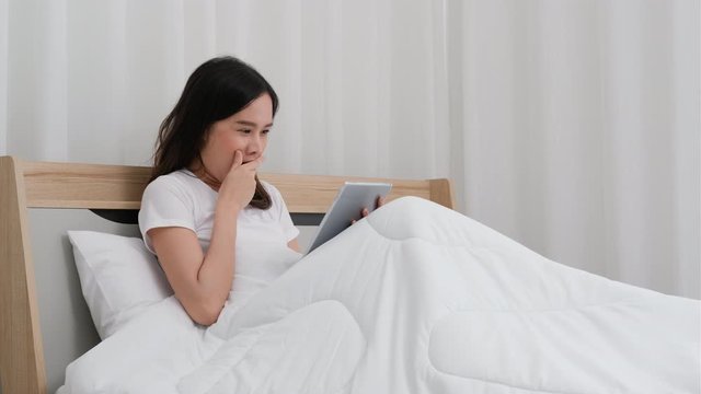 Beautiful Asian woman use tablet on bed and look sleepy by yawning then lie on white bed.