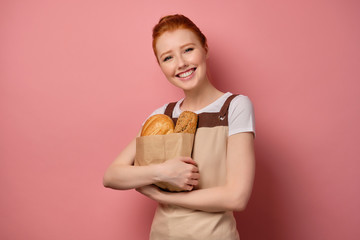 Red-haired girl with gathered hair, in an apron, stands on a pink background hugging a paper bag with bread