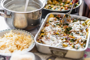 Homemade preparation of lasagna with zucchini and bacon. Close up of female hand putting and spreading bechamel and courgettes cut into small pieces on lasagna. Selective focus.Pots, plates and trays