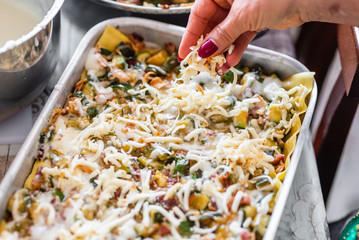 Homemade preparation of lasagna with zucchini and bacon. Close up of female hand putting and spreading bechamel and courgettes cut into small pieces on lasagna. Selective focus.Pots, plates and trays