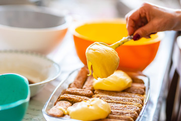 Close up of female hand putting and spreading mascarpone cream with spoon on tiramisu. Homemade preparation of italian cake tiramisu. Selective focus. Blurred terrines and bowls in the background