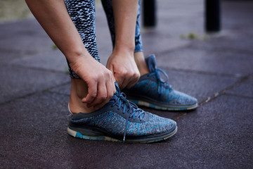 Young sportswoman, wearing grey leggings putting on sport shoes. Close-up picture of hans, showing the process of fastening shoelaces on fitness sneakers. Woman, preparing for training,
