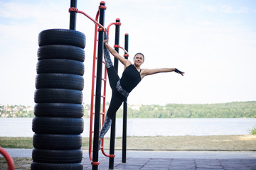Young brunette woman, wearing black fitness overall, doing stretching exercises, hanging in twine on swedish wall by city lake in summer. Fit sportswoman on outside training. Healthy lifestyle concept