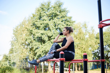 Young brunette woman, wearing black fitness overall, sitting on top of horizontal bars on sports playground in summer. Fit sportswoman outside, relaxing after training.Healthy lifestyle concept.