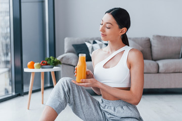 Holds glass with orange juice in hands. Young woman with slim body shape in sportswear have fitness day indoors at home