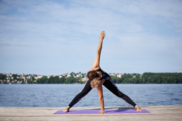Young brunette woman with bare feet, wearing black and purple fitness outfit, stretching on violet yoga mat outside on wooden pier in summer. Fit girl, doing yoga poses by lake. Healthy lifestyle.