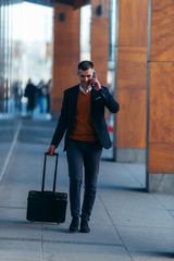 Young businessman walking through a station while talking on the phone and pulling his travel bag.