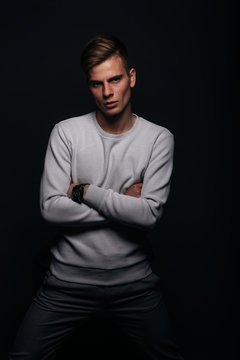 Informally ( casual ) dressed blonde young man with sharp jawline in his 20's posing in a studio in front of a black background while wearing a white sweater.