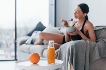 Young woman with slim body shape in sportswear sits on sofa and eats healthy diet food indoors at...