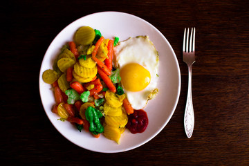 Fried egg with delicious vegetables on a white plate and a metal fork located on a dark wooden textured table.