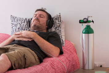 A man lies on his bed while breaths through a cannula conected to an oxygen cylinder