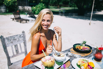 Glad adult woman eating at table on sand beach during sunny day