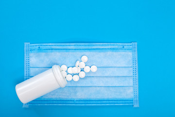 sick mask with a white plastic bottle of a medical product with a scattered pile of pills on a blue background with copy space top view.