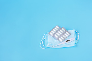  Face mask, thermometer, pills on a blue background. Medicine supplies for home coronavirus COVID-19 quarantine. Copy Space