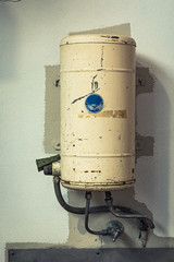 Old electric hot water service tank with pipes and taps over a sink, mounted on a painted wall in a factory.