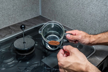 Close up over the shoulder angle of a person's hands fixing coffee in a black and stainless steal metal french press over a small stove covered in glass and grey counter top inside one early morning.