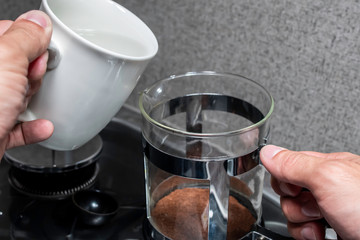 Close up over the shoulder angle of a person's hands fixing coffee in a black and stainless steal metal french press over a small stove covered in glass and grey counter top inside one early morning.