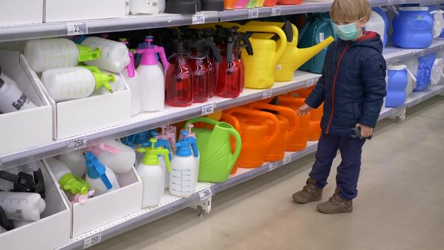 funny child carefully chooses garden watering can in supermarket. boy in medical masks because of epidemic
