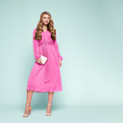 Blonde young woman in elegant pink dress. Model posing on a blue background in full length. Jewelry and hairstyle. Girl with handbag. Fashion photo