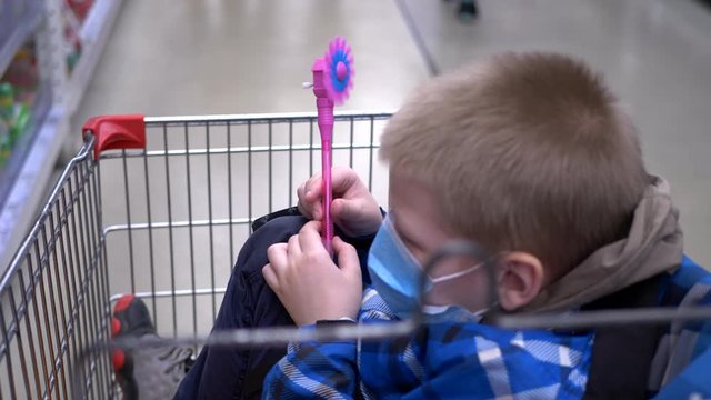 Funny boy in checkered jacket and a medical mask sits in supermarket grocery cart and plays with propeller on pencil. second child appears and fights with him toy with propeller