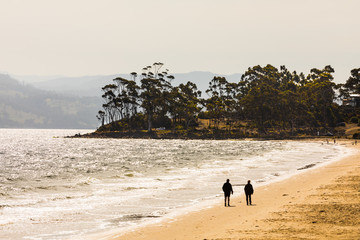 A couple walking along the curved shore of a golden sand beach on a breezy windy day towards a point.