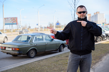 Street photo of a handsome man in a black jacket with glasses in the city on a sunny day on the road shows his fingers to the sides