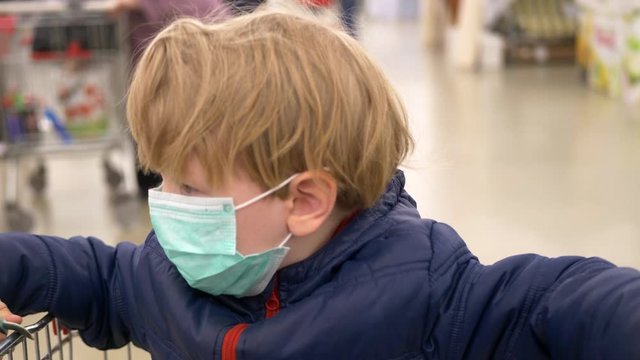 little funny blond-haired boy is sitting in supermarket grocery cart. child was put on protective medical mask from coronavirus, which he is trying to remove. Infection protection concept