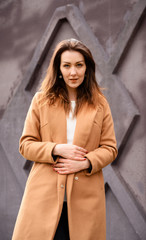 Portrait of a brunette girl standing in spring in a beige coat walk a street on a background of a dark purple wall of a building