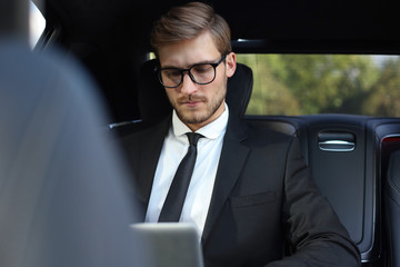 Handsome confident businessman in suit talking on smart phone and working using laptop while sitting in the car.