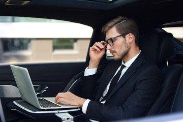 Thoughtful confident businessman keeping hand on glasses while sitting in the luxe car and using his laptop.