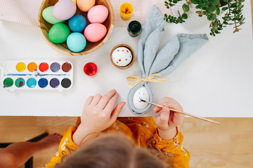 Little girl painting easter eggs behind a table using brush. Top view.