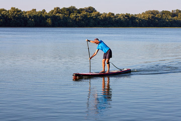 Naklejka premium Joyful man is training on a SUP board on Danube river during sunny morning. Stand up paddle boarding - awesome active recreation in nature