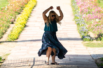 Blonde woman dances wearing a flowing long maxi skirt on a pathway lined with flowers. Taken in Lodi Gardens New Delhi India