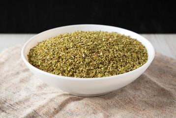 Dried Green Greek Oregano Spice in a white bowl on cloth, low angle view. Close-up.