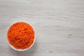 Fototapeta na wymiar Shredded carrots in a white bowl on a white wooden background, top view. Flat lay, overhead, from above. Copy space.