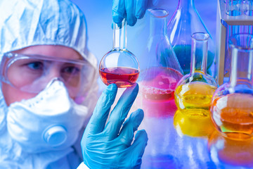 A chemist examines a substance in a flask against a background of chemical dishes with colored chemical liquids. Analysis of chemical samples. Adding various reagents to the test substance.