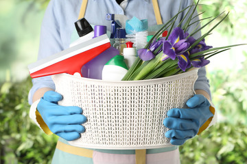 Woman holding basket with spring flowers and cleaning supplies on green blurred background, closeup