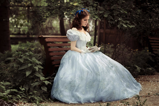 A young beautiful girl in a full blue dress, with curly hair, is sitting on a bench, holding a book in her hands and reading it. Against a tree.Full-length photo. Historical reconstruction